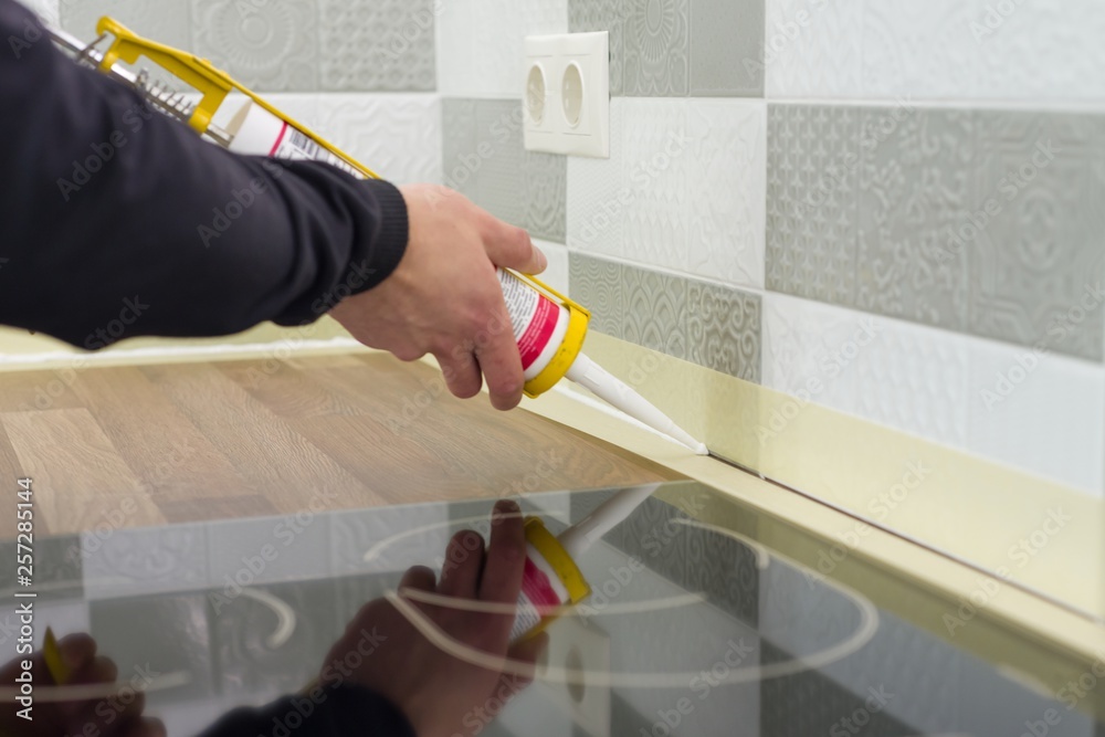 Applying silicone sealant with construction syringe. Worker fills seam between the ceramic tiles on the wall and kitchen worktop - obrazy, fototapety, plakaty 