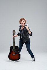 Wall Mural - Young woman musician in a black leather jacket with an acoustic guitar in hand on a gray background. He laughs and plays rock and roll loudly. Full-length portrait