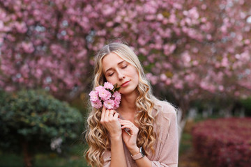 Beautiful young woman with long curly blonde hair and closed eyes holding blooming branch of sakura tree