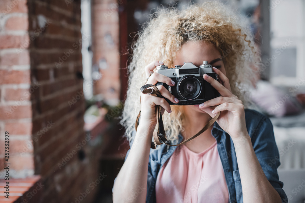 Curly blonde woman holding her retro photo camera at home. - obrazy, fototapety, plakaty 