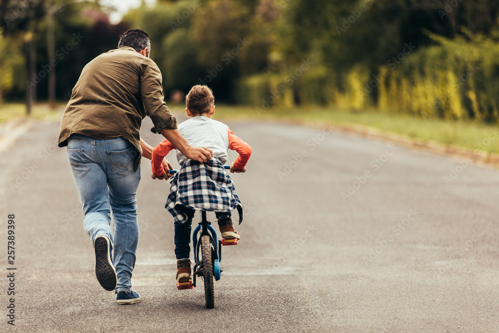 Man helping his kid in learning to ride a bicycle - obrazy, fototapety, plakaty 