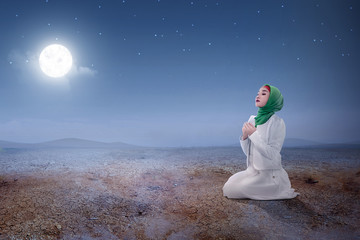 Wall Mural - Young asian muslim woman sitting in pray position while raised hands and praying on the sand dune