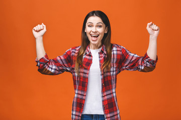 Photo of cheerful beautiful young woman standing isolated over orange wall background. Showing winner gesture.