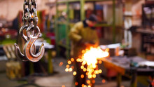 Construction Plant. A Big Industrial Lifting Chain With A Hook On The End Hanging In The Air. A Man Welding On The Background