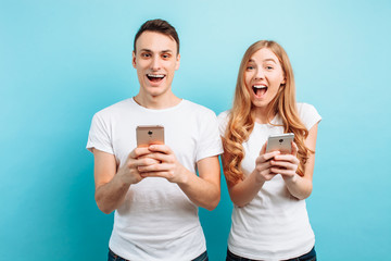 Photo of excited people, man and woman, rejoicing, holding smartphones in their hands, on light blue background