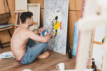 Selective focus of half-naked artist in blue jeans sitting on floor and painting picture on canvas