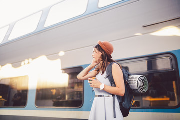 theme tourism and travel young student. beautiful young Caucasian girl in dress and hat standing at train station near train with backpack and cup coffee in hand. Uses smartphone phone technology