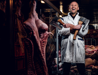 Cheerful butcher in workwear posing with two axes in a refrigerated warehouse in the midst of meat carcasses