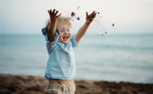 A Small Toddler Boy Standing On Beach On Summer Holiday, Throwing Sand.