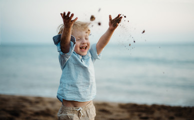 a small toddler boy standing on beach on summer holiday, throwing sand.