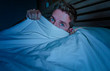 © TheVisualsYouNeed - portrait of young attractive scared man in fear and panic suffering horror nightmare covering face with blanket sleepless at night lying on bed