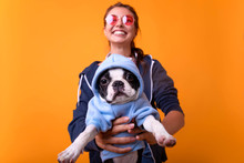 Young Hipster Smiling Woman Hugging Her Pet At Yellow Background.Cheerful Young Woman In Trendy Sportswear Holding Her Big Puppy And Laughing. Indoor Close Up Portrait