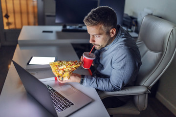 Young Caucasian architect drinking juice and eating pizza at office. On desk laptop. Late night work concept.