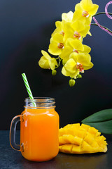 Wall Mural - Mango smoothie in a glass jar and fresh mango on a black background. Mango shake. Tropical fruit concept.