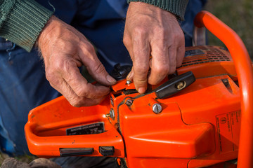 Canvas Print - Old man in blue pants repair orange chainsaw placed on the ground with his bare hands.