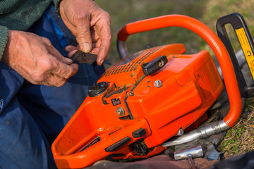 Canvas Print - Old man in blue pants repair orange chainsaw placed on the ground with his bare hands.