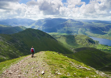 A Female Hiker And Their Dog Descending From Grasmoor Down Lad Hows Above Buttermere On A Sunny Day In The English Lake District, UK.