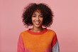 Indoor portrait of positive optimistic charming african american girl with afro hairstyle looks with pleasure, with lively smile, wearing colorful longsleeve, isolated on pink background