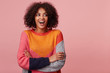 Lively charismatic attractive African American with an afro hairstyle with excitement looks left to the empty space, laughs, ha-ha, stand with arms crossed, wearing colorful sweater, isolated on pink