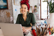 © Anton - Young attractive woman with dark curly hair sitting at the table with laptop dreamily looking in camera while spending time in modern cozy workshop with big windows