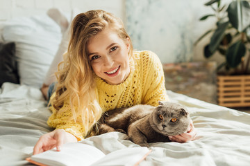 beautiful young woman looking at camera and holding book while lying in bed with scottish fold cat