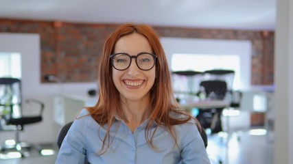 Poster - employee with red long hair and eyeglasses laughing looking at the camera. portrait young caucasian businesswoman sitting at the workplace in modern office.