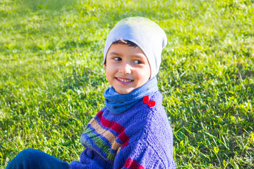 Wall Mural - cheerful boy in a hat and sweater sitting on the grass