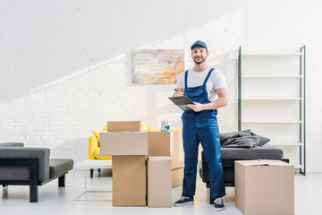 Wall Mural - mover looking at camera and holding clipboard near cardboard boxes in apartment with copy space