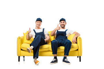 two movers in uniform sitting on yellow sofa, showing thumb up signs and looking at camera isolated on white