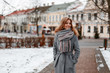 © alones - Urban pretty young woman with a beautiful smile in a gray stylish coat in a knitted warm scarf walks around the city near the vintage buildings on a winter day. Attractive happy girl on vacation.