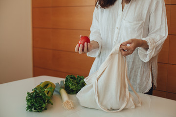 Hipster girl putting greens and fresh leek on the kitchen table out of the cotton reusable tote bag, using eco shopper instead of a plastic bag, concept of healthy lifestyle and saving the planet