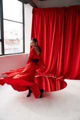 Wall Mural - Sexy brunette woman in long red dress dancing in studio with large windows and red curtains.