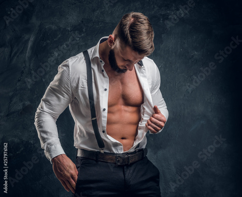 Fototapeta na szkło A stylish young man with a muscular body in the unbuttoned white shirt with suspenders. Studio photo against a dark wall background