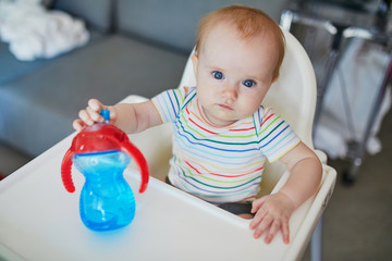 Wall Mural - Little baby girl sitting in high chair and drinking water from sippy cup