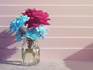 Spring composition. A bouquet of large red roses and several small blue chrysanthemums is in a glass mug. Light wooden background.