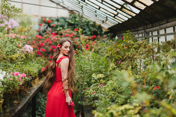 Young beautiful caucasian woman in glass greenhouse among colorful azalea flowers. Art portrait of a long-haired girl wearing a romantic red dress.