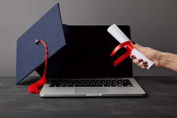 Poster - Cropped view of woman holding diploma near laptop with blank screen and academic cap on grey