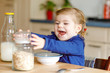 Adorable toddler girl eating healthy oatmeals with milk for breakfast. Cute happy baby child in colorful clothes sitting in kitchen and having fun with preparing oats, cereals. Indoors at home
