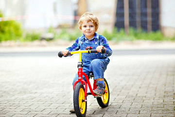 Active blond kid boy in colorful clothes driving balance and learner's bike or bicycle in domestic garden. Toddler child dreaming and having fun on warm summer day. outdoors movement game for children