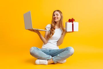 Portrait of an excited girl sitting on the floor with a laptop and a gift box in her hands, on a yellow background
