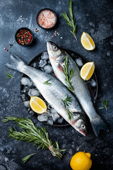 Wall Mural - Fresh raw seabass fish on black stone background with spices, herbs, lemon and salt. Culinary seafood background with ingredients for cooking. Top view