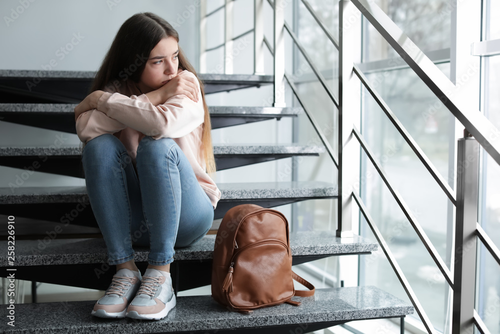 Upset teenage girl with backpack sitting on stairs indoors. Space for text - obrazy, fototapety, plakaty 