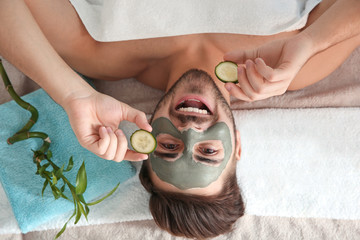 Sticker - Young man with clay mask on his face holding cucumber slices in spa salon, above view