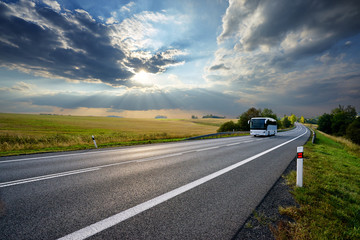 Wall Mural - white bus traveling on the asphalt road in rural landscape at sunset with dramatic clouds