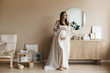 Beautiful brown-haired pregnant model girl with blue eyes and with a pretty smile in a stylish beige dress posing at minimalist interior
