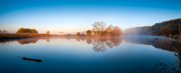 Wall Mural - calm lake in autumn at sunrise, fog rising from the water, blue sky