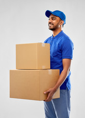 Wall Mural - mail service and shipment concept - happy indian delivery man with parcel boxes in blue uniform over grey background