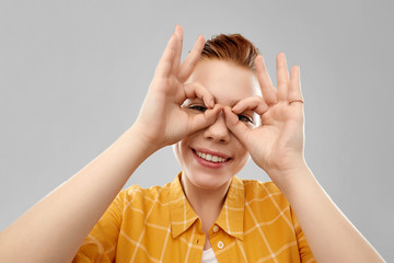 people concept - smiling red haired teenage girl in checkered shirt looking through finger glasses over grey background