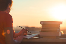 Female Student With A Lot Of Books Preparing For University Exam Late Evening On The Sunset Town Background. Education Concept