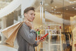 © mad_production - Young handsome man using his smart phone at the shopping mall, copy space. CHeerful male customer enjoying seasonal sale at the mall, carrying shopping bags with purchase. Social media, online concept
